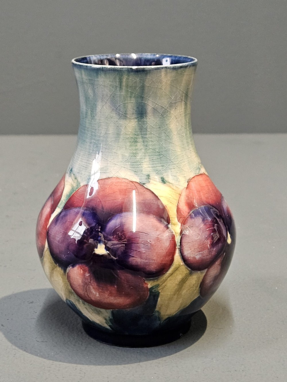 A Moorcroft earthenware vase featuring a bulbous body and flared neck decorated with hand-painted tubelined purple and wine-red pansies beneath a pale blue glaze. View V