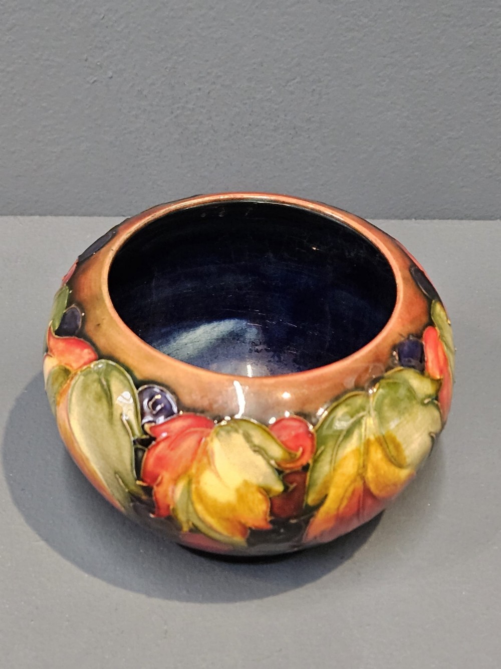 A small round earthenware vase decorated with tubelined autumn leaves and berries in yellow, green, and red against a dark flambe glaze. View II