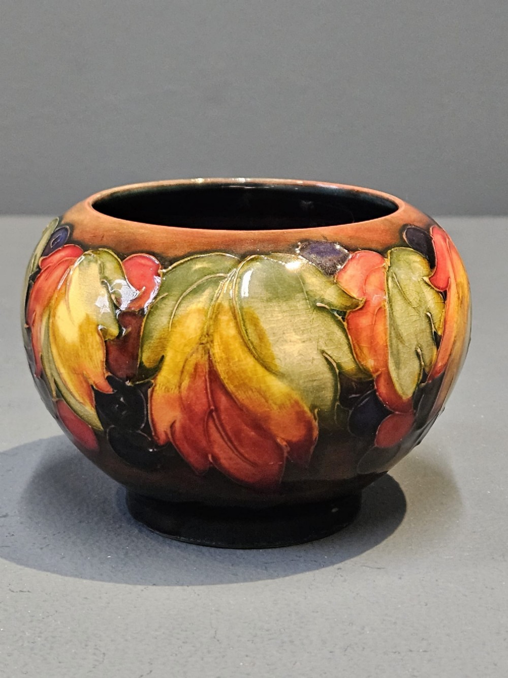 A small round earthenware vase decorated with tubelined autumn leaves and berries in yellow, green, and red against a dark flambe glaze. View V