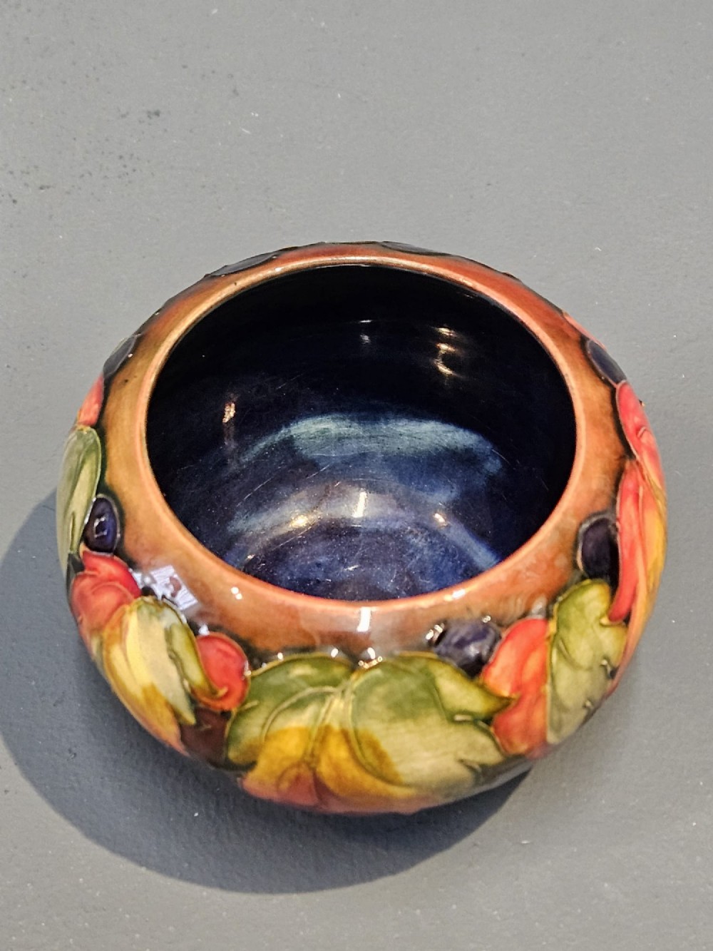 A small round earthenware vase decorated with tubelined autumn leaves and berries in yellow, green, and red against a dark flambe glaze. View VI