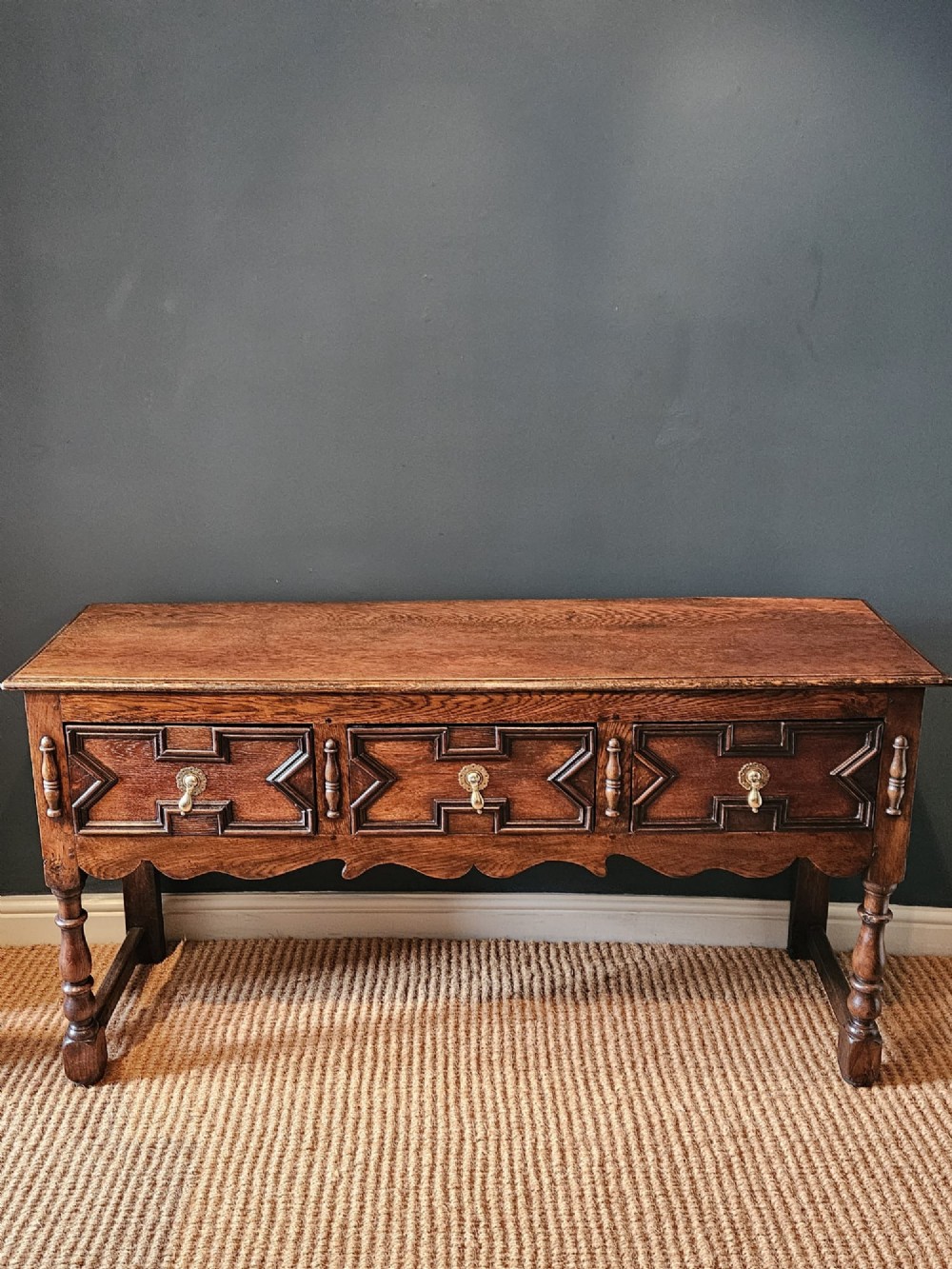 An early 18th-century George I oak dresser featuring three drawers with geometric mitred mouldings, brass drop handles, a scalloped apron, and turned legs.