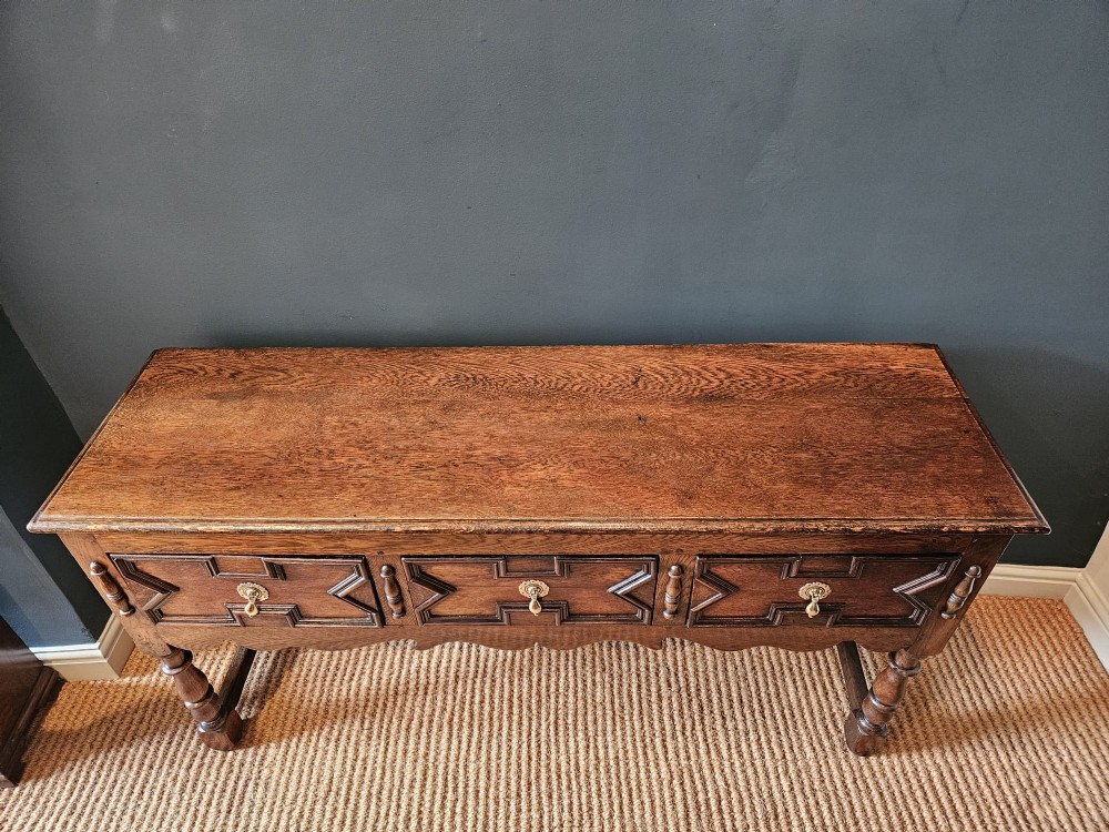 An early 18th-century George I oak dresser featuring three drawers with geometric mitred mouldings, brass drop handles, a scalloped apron, and turned legs. View VIII