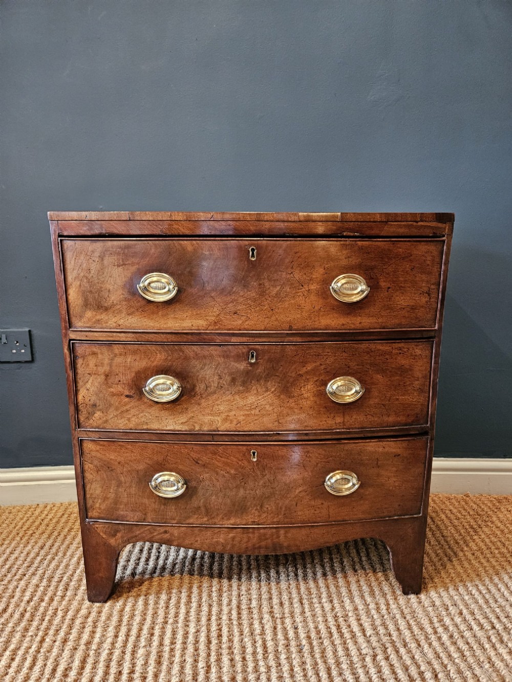 An early Victorian mahogany bow-fronted chest featuring three full-length drawers with oval brass handles and a curved lower apron. View II