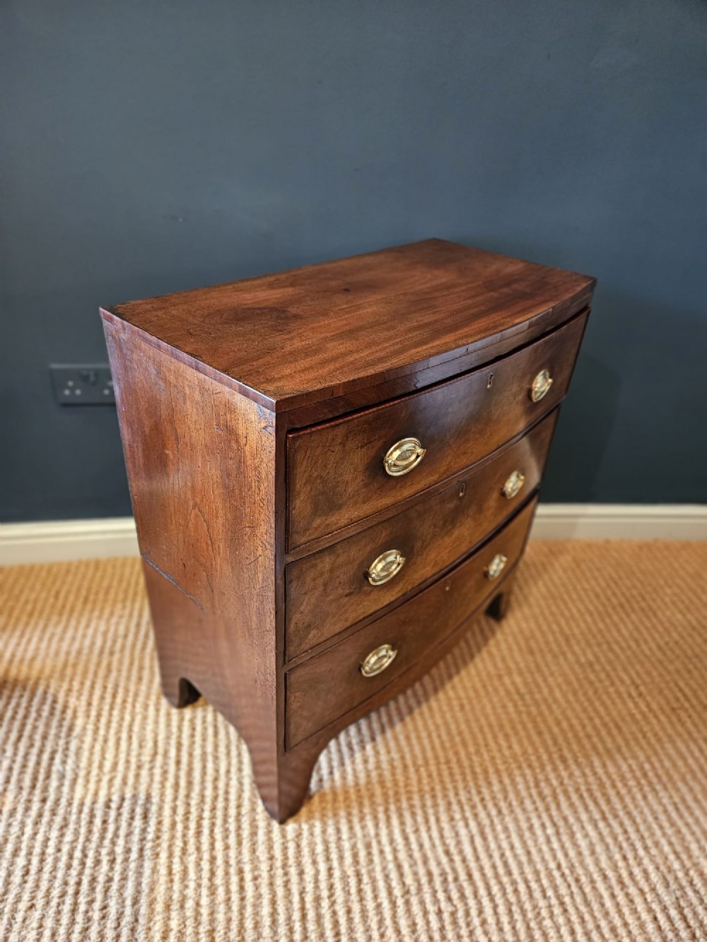 An early Victorian mahogany bow-fronted chest featuring three full-length drawers with oval brass handles and a curved lower apron. View V