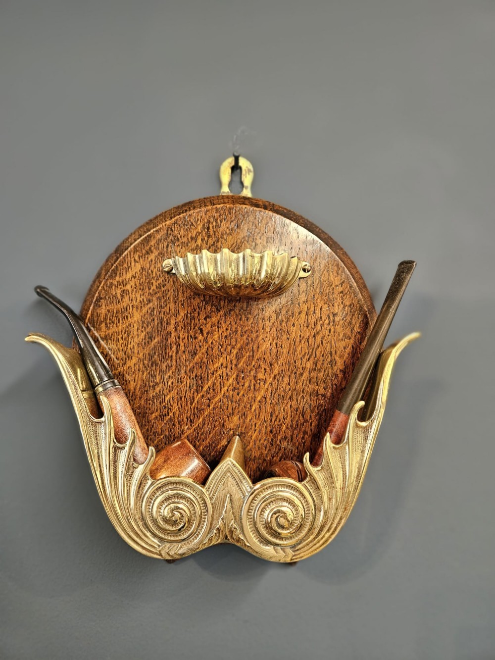 A Victorian wall-mounted oak pipe rack featuring ornamental cast brass scrollwork holders and a brass shell motif.