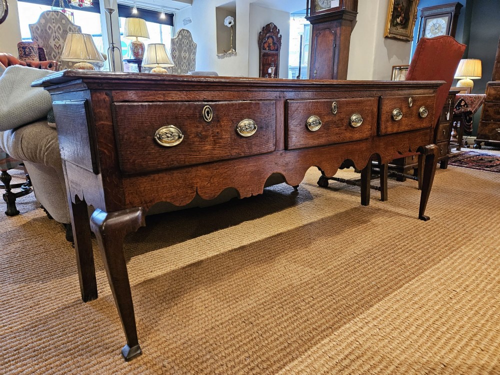 A George II oak dresser base featuring three drawers, brass handles, a shaped apron, and four cabriole legs.