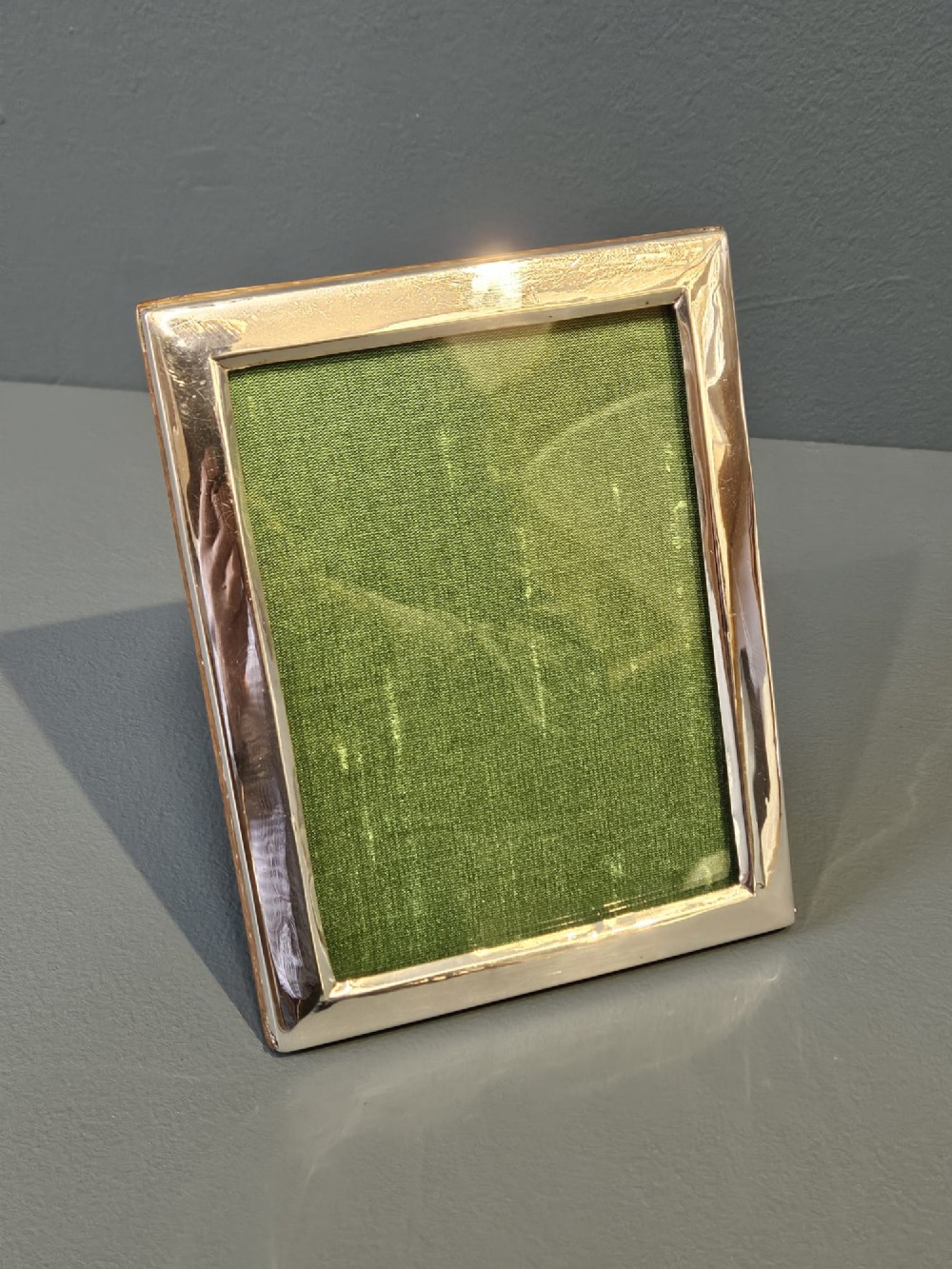 A rectangular George V silver photo frame with a plain polished border and a green fabric insert resting on a neutral surface.