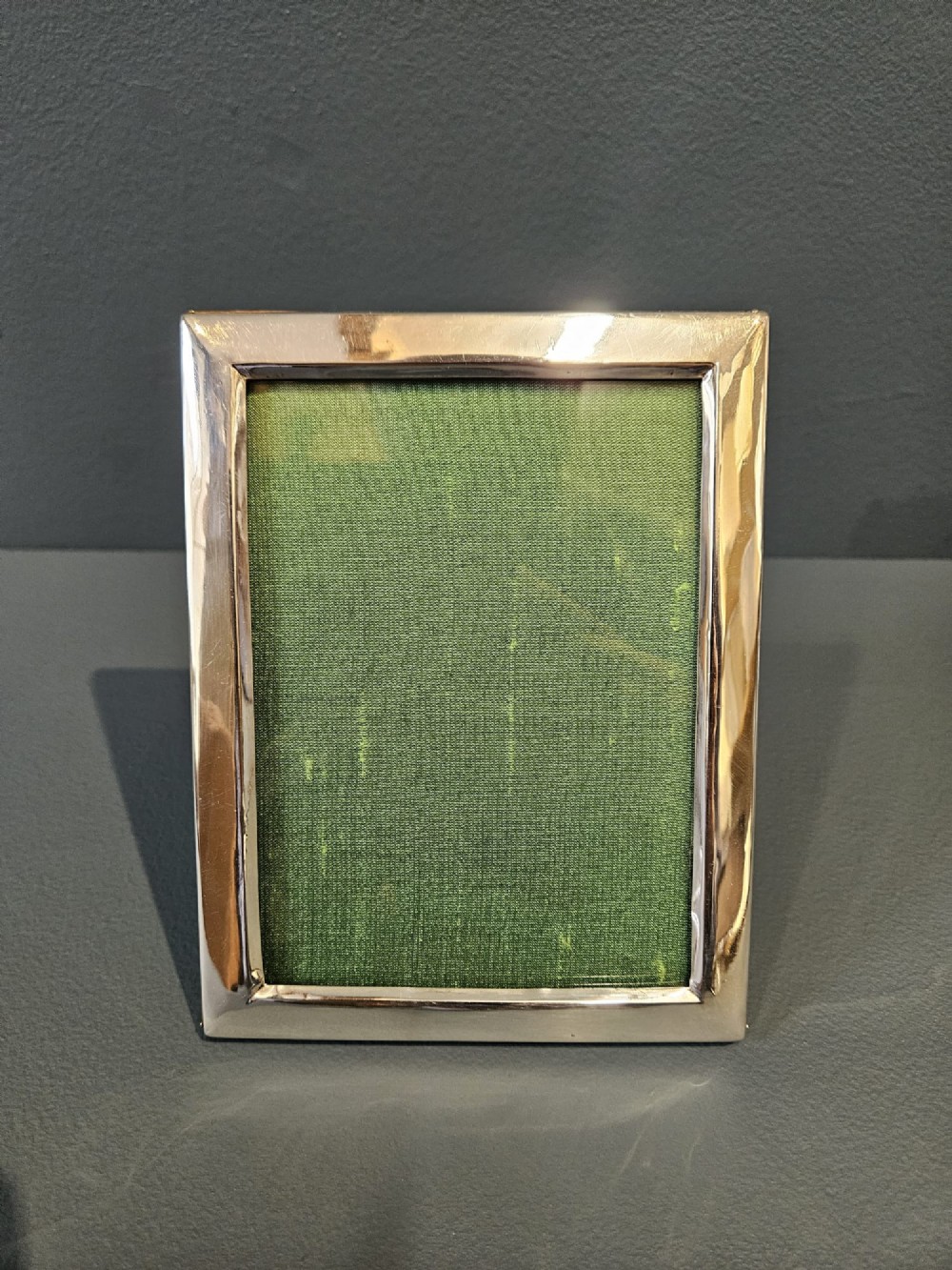 A rectangular George V silver photo frame with a plain polished border and a green fabric insert resting on a neutral surface. View IV