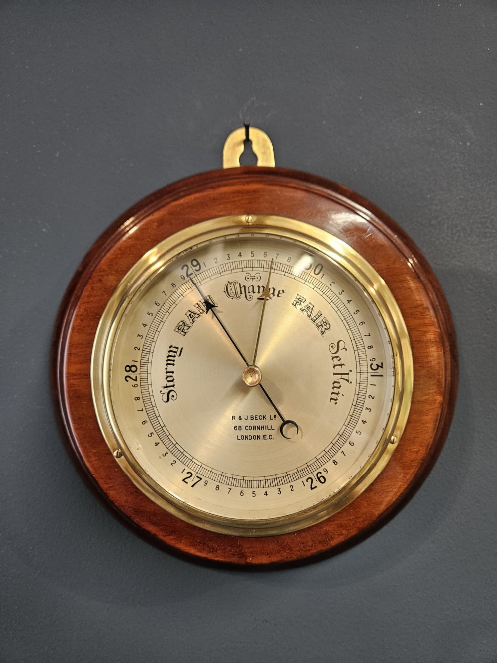 A circular Victorian aneroid barometer set within a polished mahogany surround with an engraved brass dial face and brass bezel.