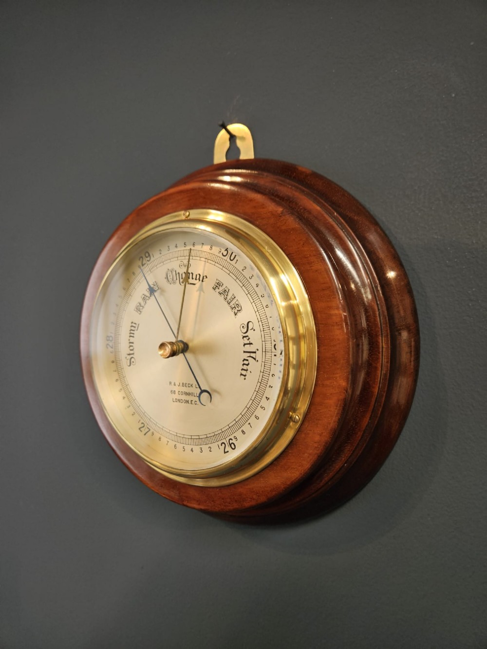 A circular Victorian aneroid barometer set within a polished mahogany surround with an engraved brass dial face and brass bezel. View II