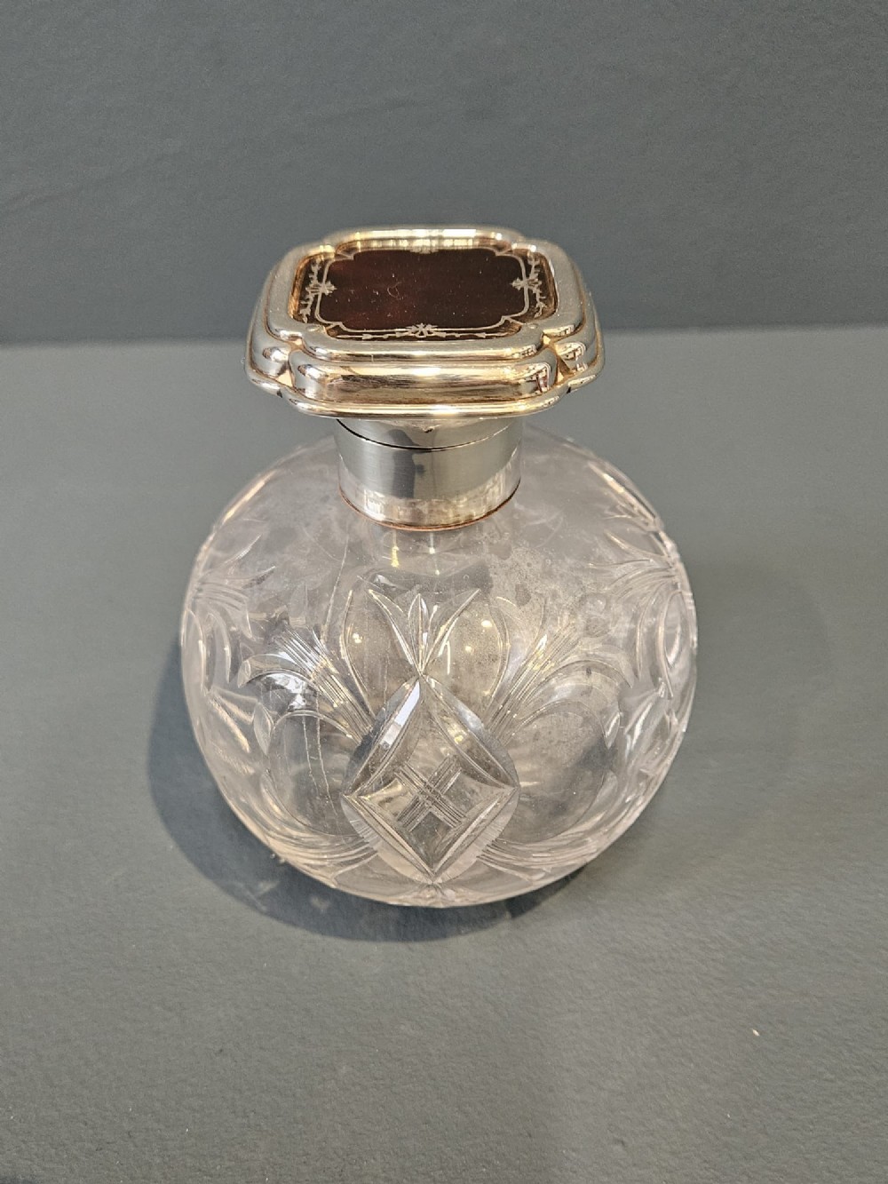 A round cut crystal glass scent bottle with a sterling silver neck collar and a square hinged silver lid featuring an inlaid tortoiseshell top.