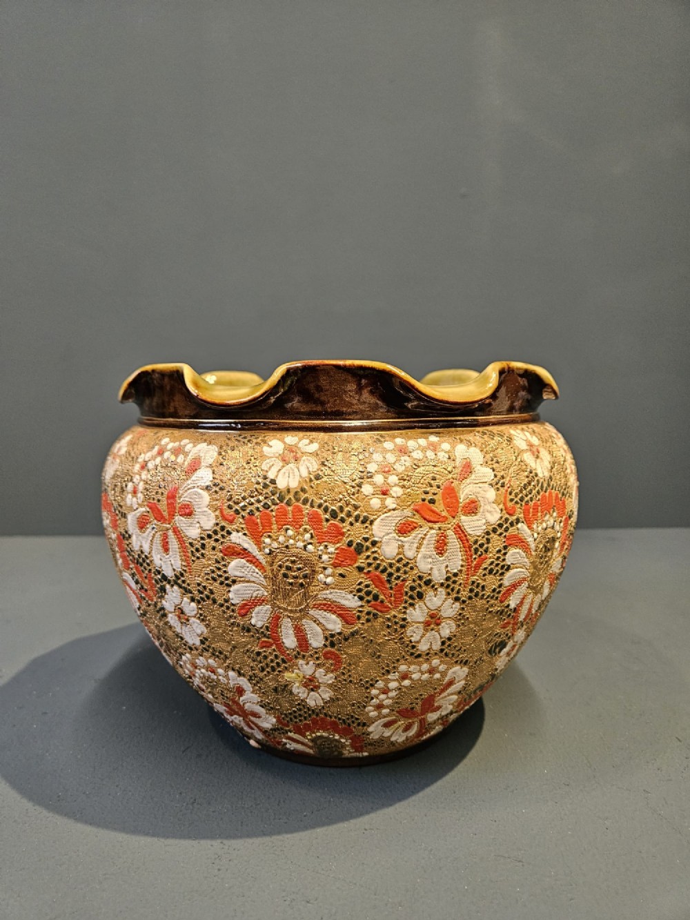A Victorian Doulton Lambeth stoneware jardiniere featuring a wavy crimped rim, textured lace-impressed floral pattern in white and terracotta red on an ochre ground, and a dark brown upper band. View V