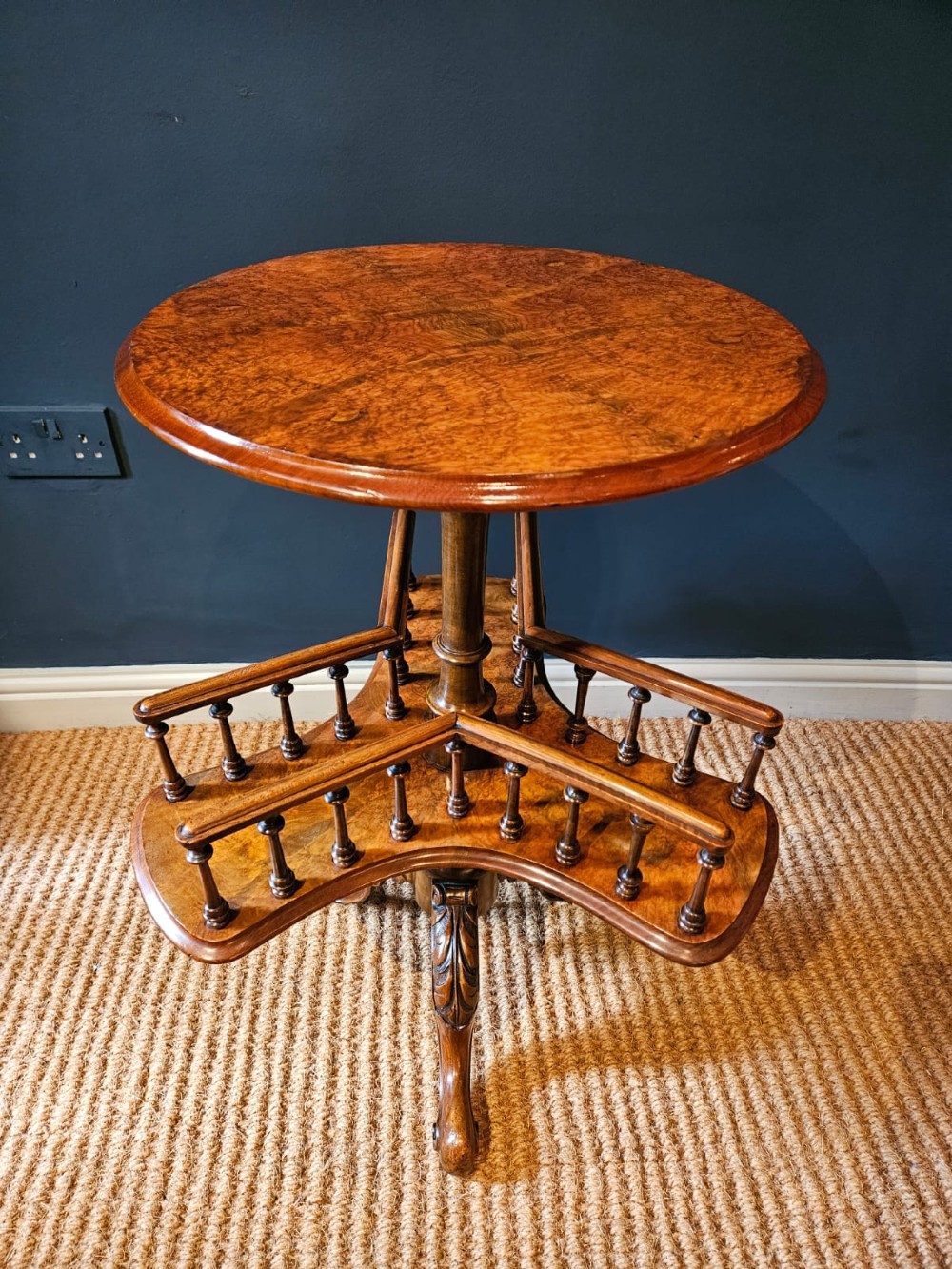 A Victorian burr walnut revolving book table with a round top, three spindle-railed book compartments, a turned central stem, and carved tripod legs.