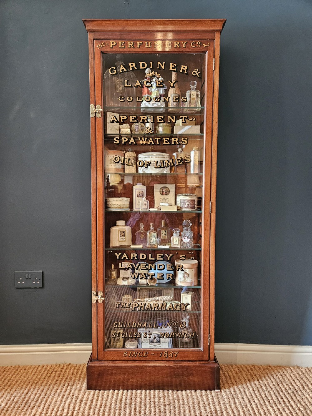 A tall Edwardian mahogany glazed display cabinet featuring gilded commercial typography on the glass door and shelves filled with apothecary bottles and packages.