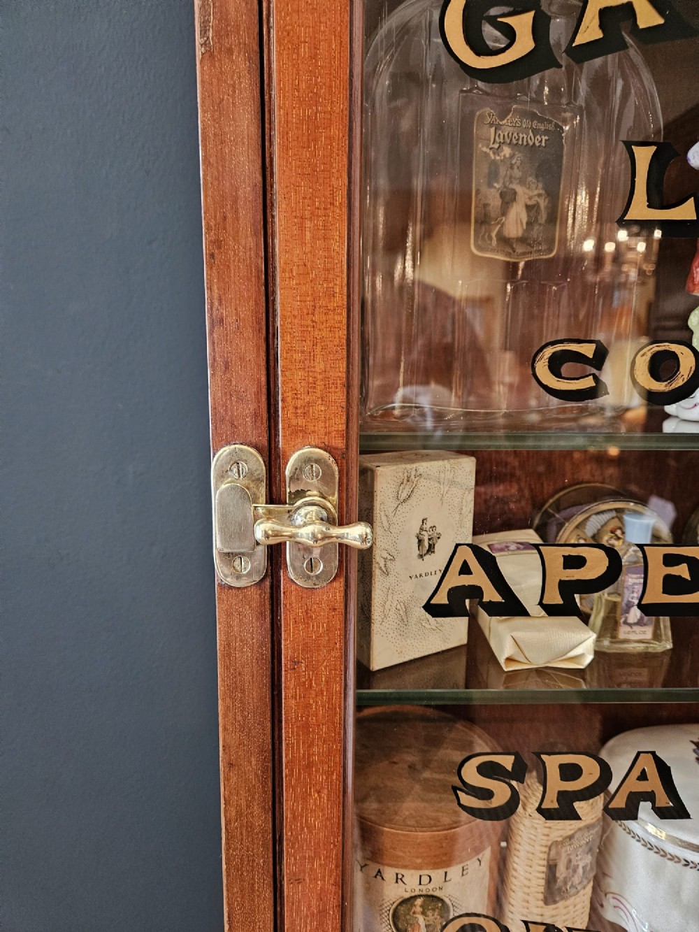 A tall Edwardian mahogany glazed display cabinet featuring gilded commercial typography on the glass door and shelves filled with apothecary bottles and packages. View II