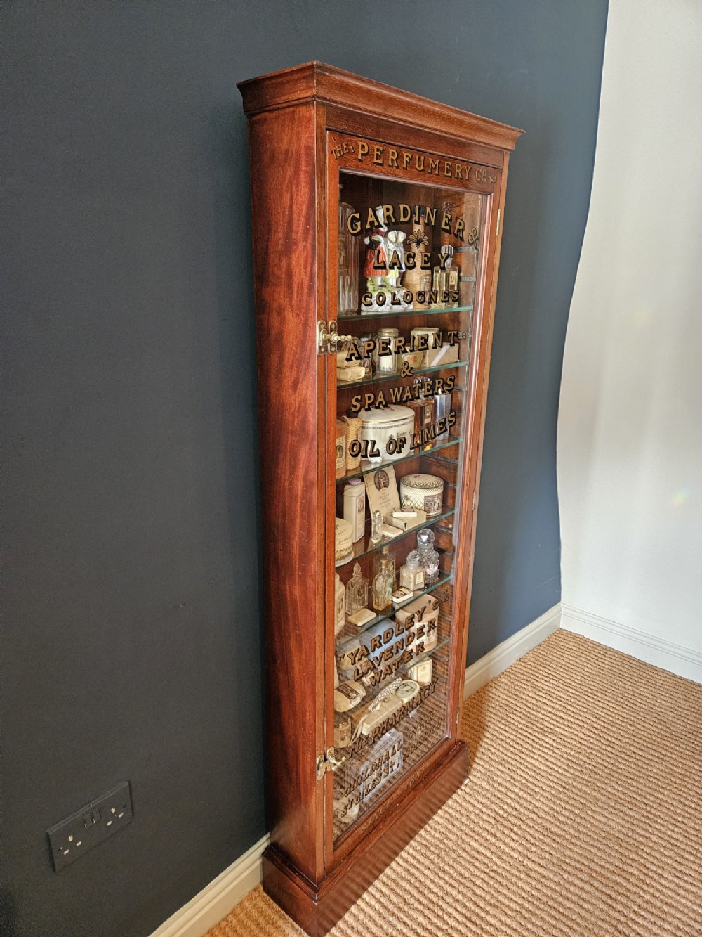 A tall Edwardian mahogany glazed display cabinet featuring gilded commercial typography on the glass door and shelves filled with apothecary bottles and packages. View III