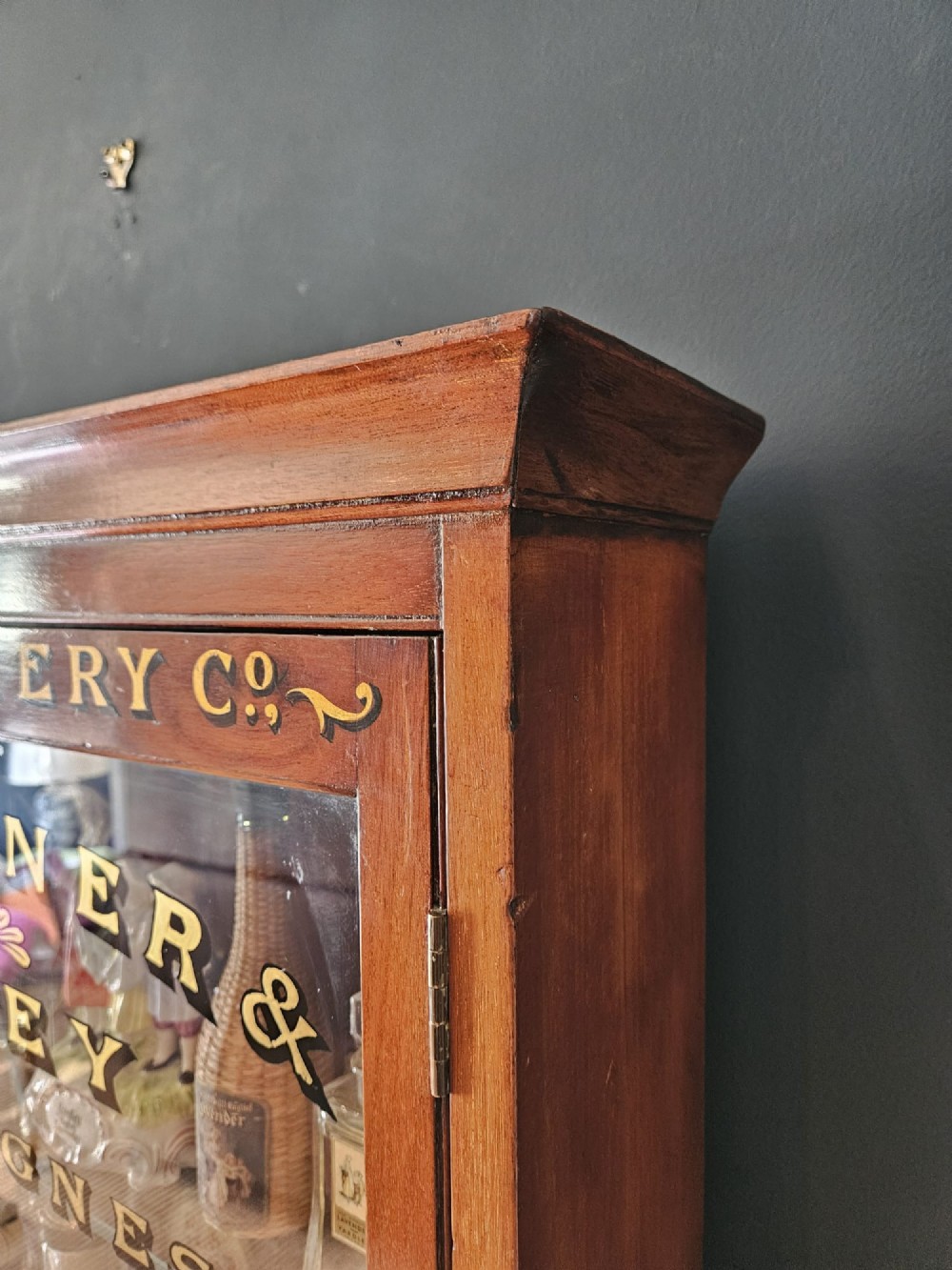 A tall Edwardian mahogany glazed display cabinet featuring gilded commercial typography on the glass door and shelves filled with apothecary bottles and packages. View V