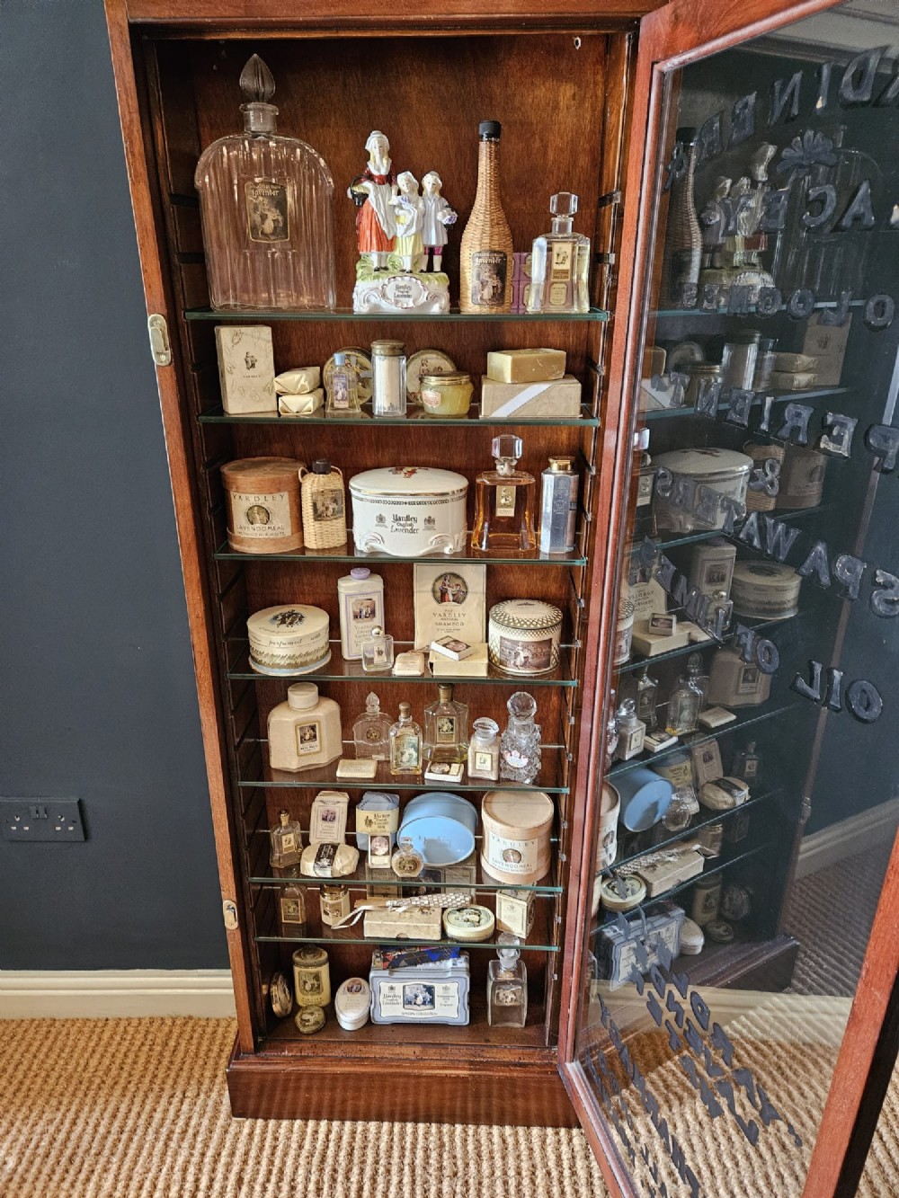 A tall Edwardian mahogany glazed display cabinet featuring gilded commercial typography on the glass door and shelves filled with apothecary bottles and packages. View VI