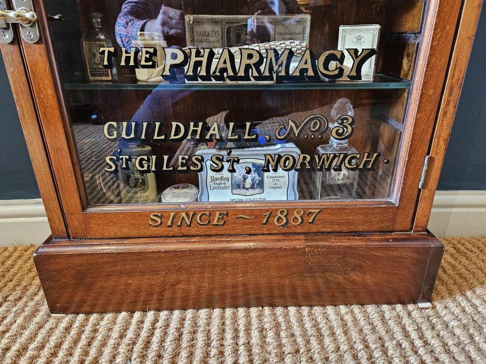A tall Edwardian mahogany glazed display cabinet featuring gilded commercial typography on the glass door and shelves filled with apothecary bottles and packages. View VIII