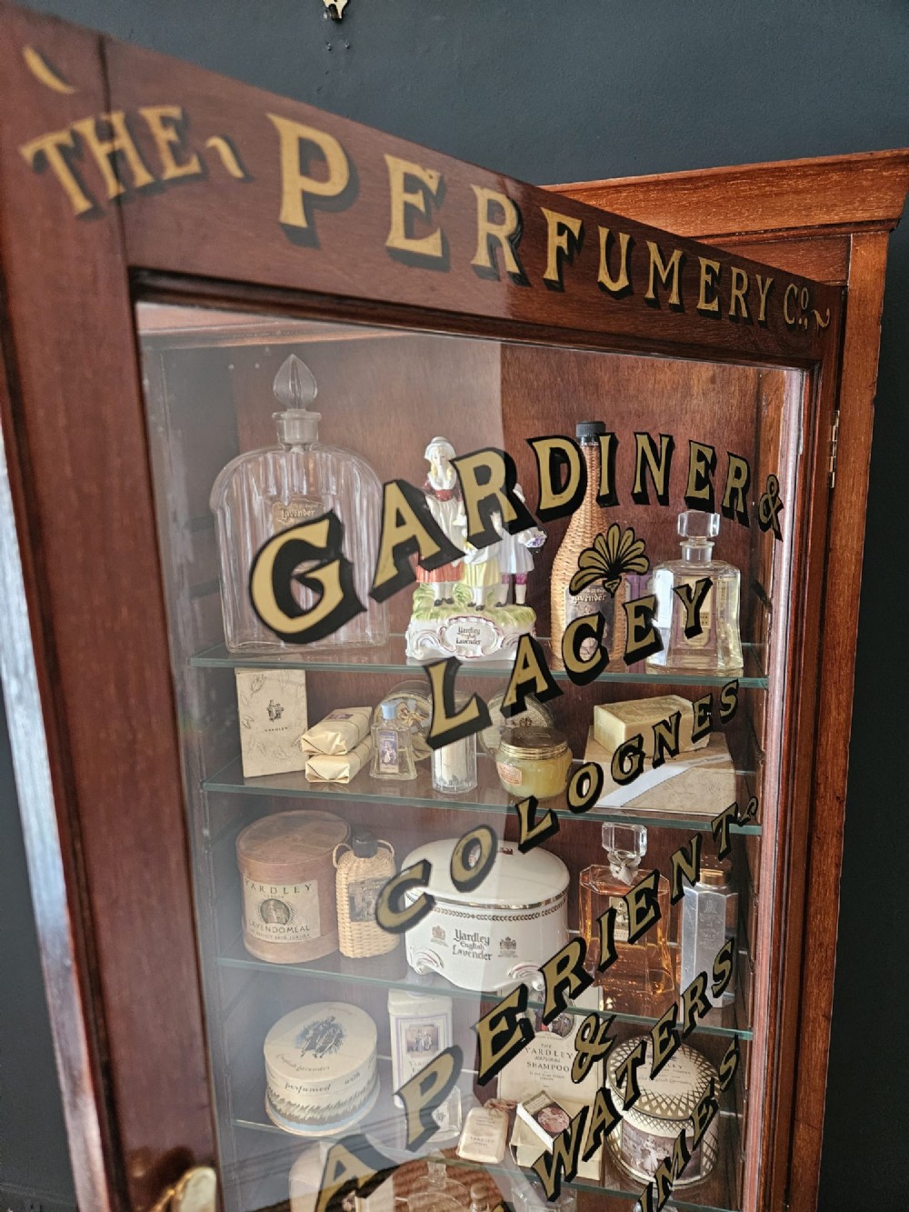 A tall Edwardian mahogany glazed display cabinet featuring gilded commercial typography on the glass door and shelves filled with apothecary bottles and packages. View IX