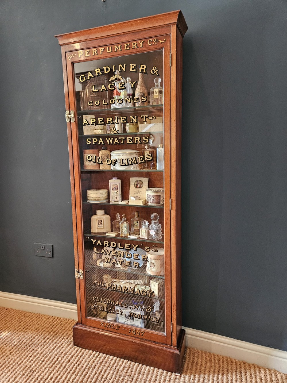 A tall Edwardian mahogany glazed display cabinet featuring gilded commercial typography on the glass door and shelves filled with apothecary bottles and packages. View X