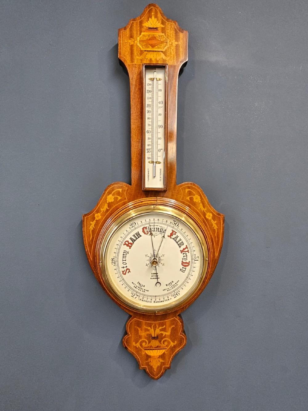 An Edwardian mahogany banjo barometer featuring decorative satinwood urn inlays, an upper thermometer gauge, and a circular aneroid pressure dial. View VI