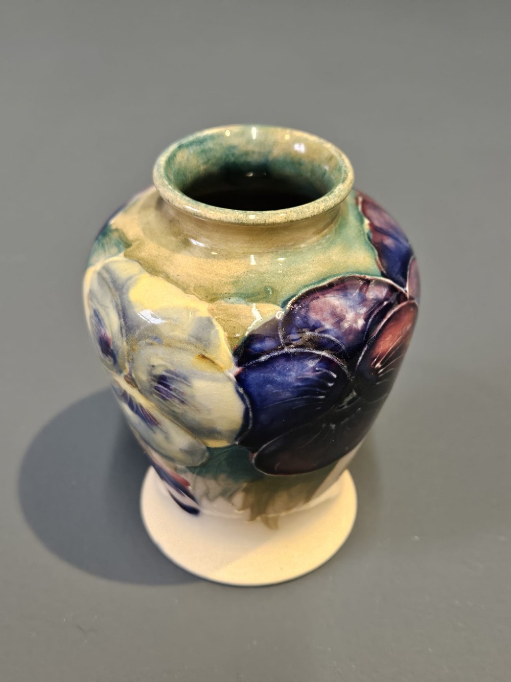 A small baluster-shaped ceramic vase decorated with hand-painted purple and blue pansies on a cream background with a flared green rim. View II