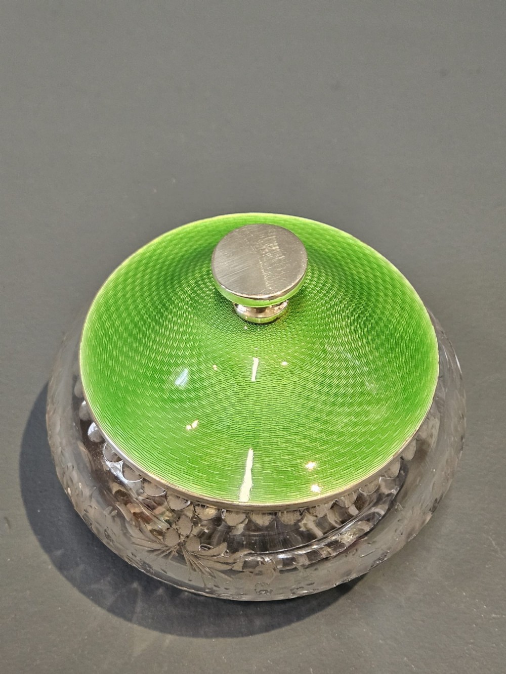 A clear cut glass round jar with a silver-rimmed lid covered in bright green engine-turned enamel and finished with a silver finial. View V