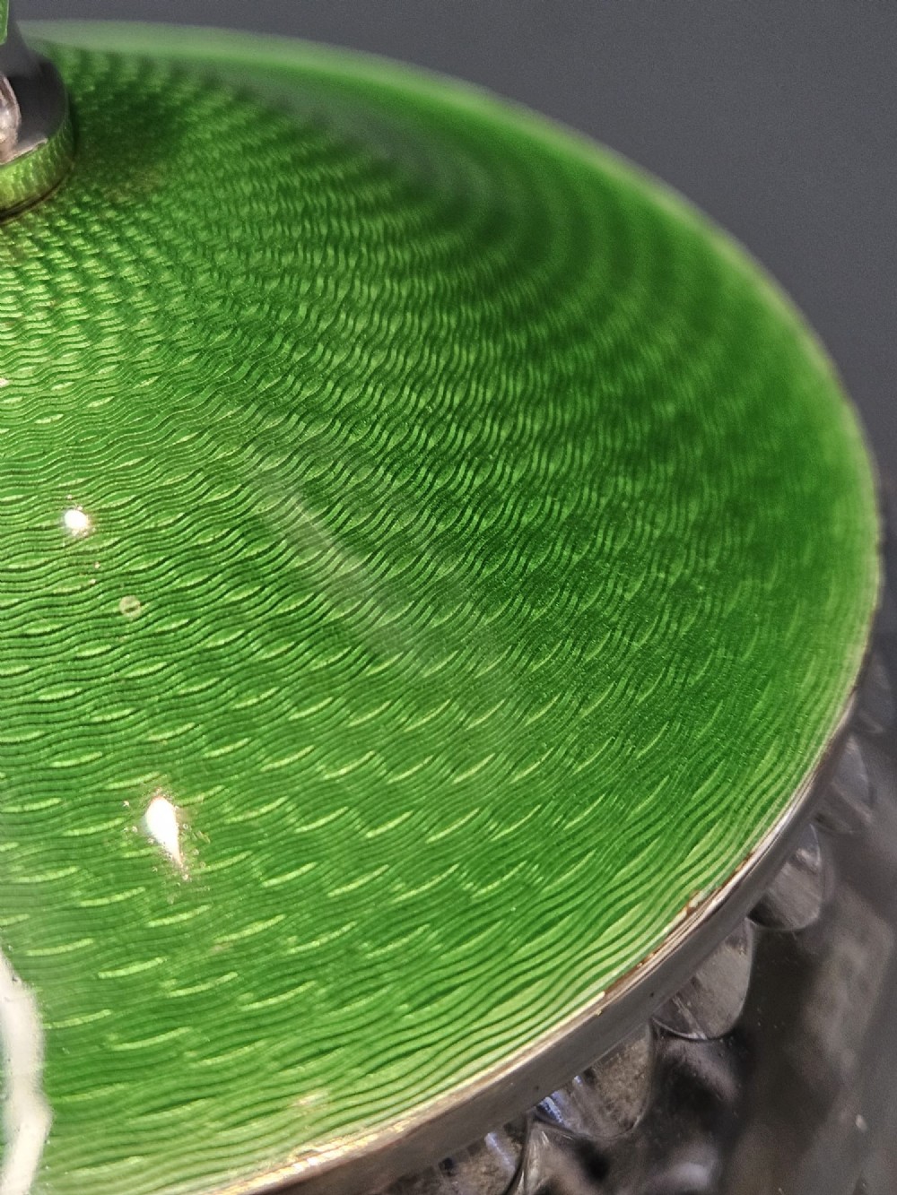 A clear cut glass round jar with a silver-rimmed lid covered in bright green engine-turned enamel and finished with a silver finial. View VI