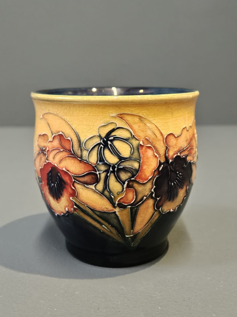 A rounded glazed ceramic vase featuring a yellow upper section decorated with tubelined orchids above a dark cobalt blue base. View II