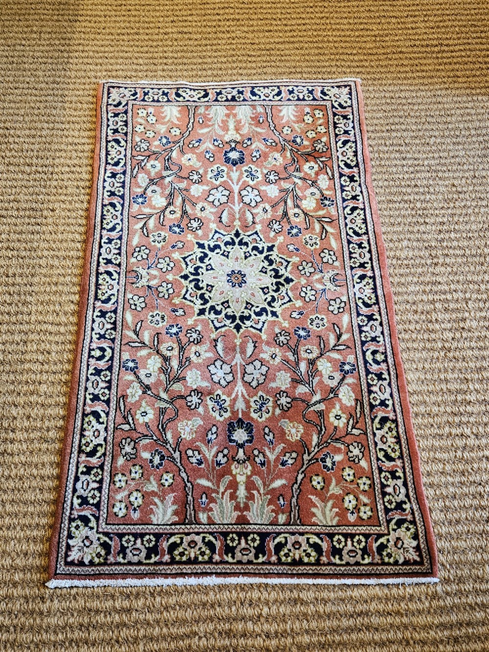 A rectangular pure wool hearth rug with a central floral medallion, salmon terracotta ground, and navy blue patterned borders. View V