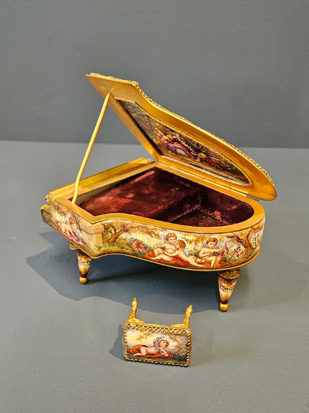 A miniature grand piano and matching stool music box crafted from gilt metal with hand-painted enamel panels depicting classical figures. View V
