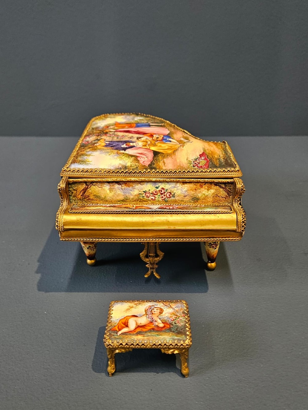 A miniature grand piano and matching stool music box crafted from gilt metal with hand-painted enamel panels depicting classical figures. View VI