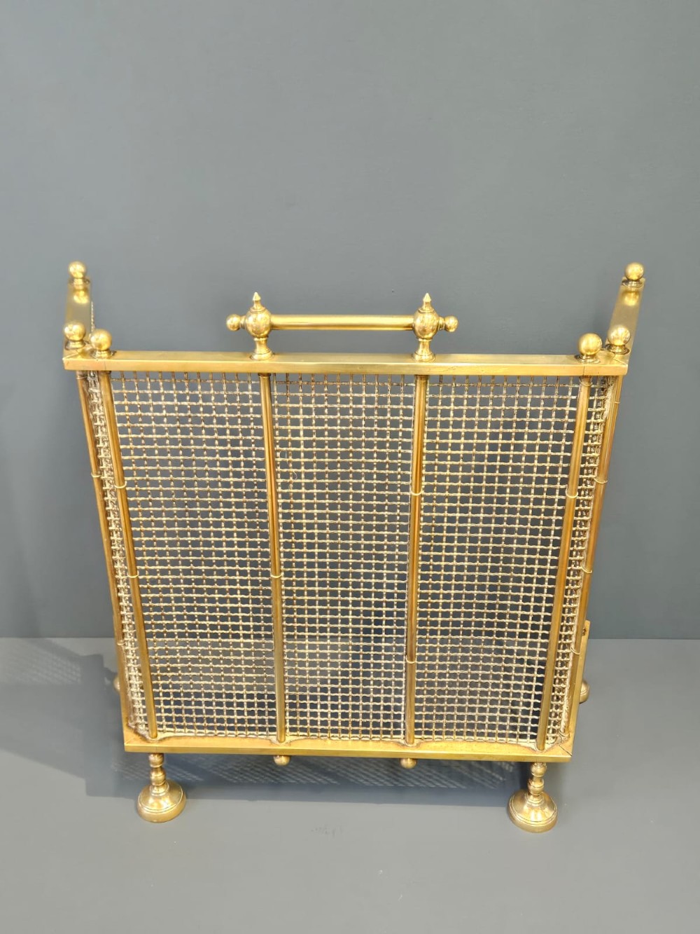 A Victorian cast brass fire guard featuring a woven wire mesh screen, turned brass finials, a central carrying handle, and small turned brass feet. View II