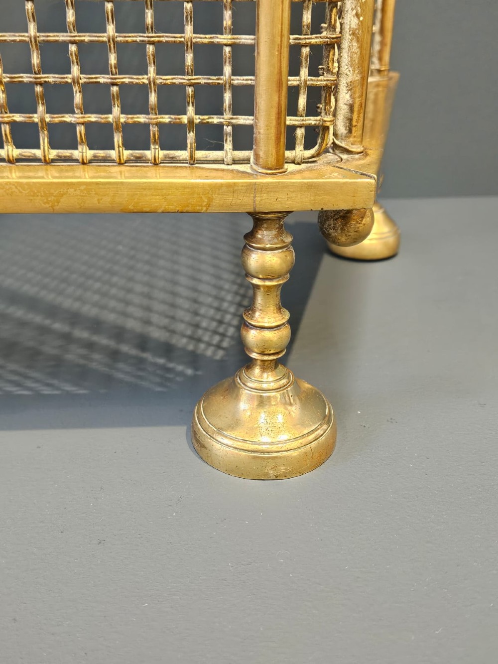 A Victorian cast brass fire guard featuring a woven wire mesh screen, turned brass finials, a central carrying handle, and small turned brass feet. View VI