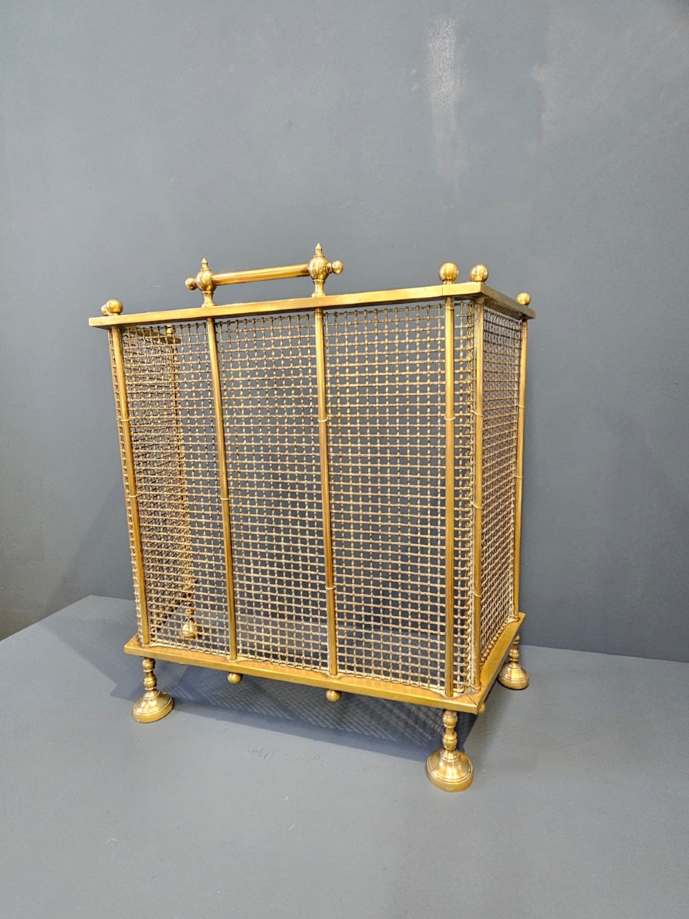 A Victorian cast brass fire guard featuring a woven wire mesh screen, turned brass finials, a central carrying handle, and small turned brass feet. View VII
