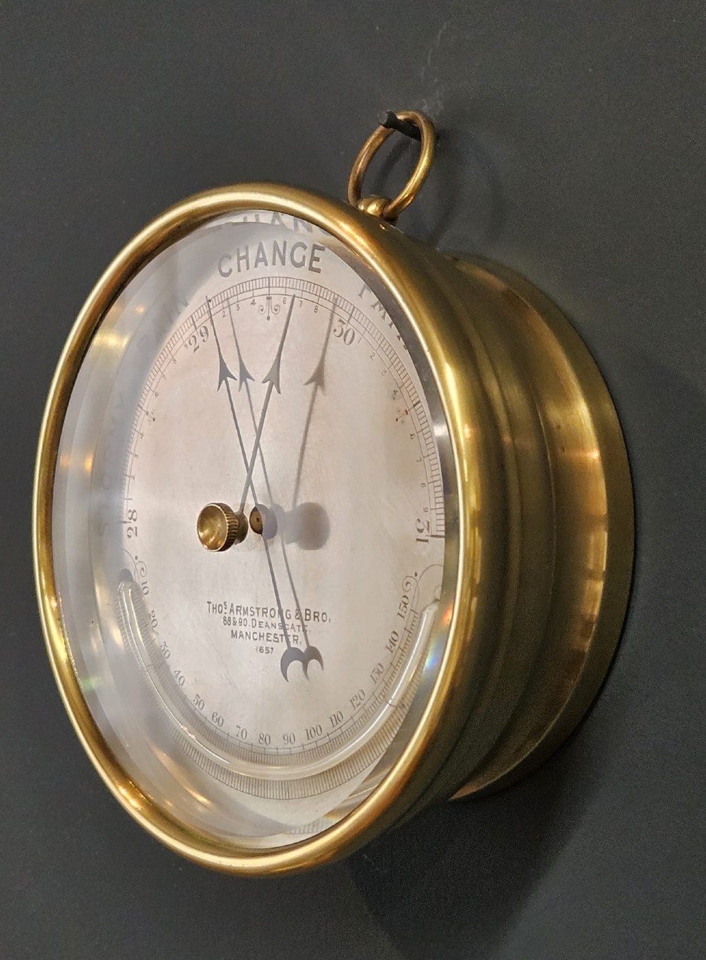 A circular Victorian brass aneroid barometer featuring an engraved silvered dial with weather scale, integrated thermometer, and a top suspension ring. View V