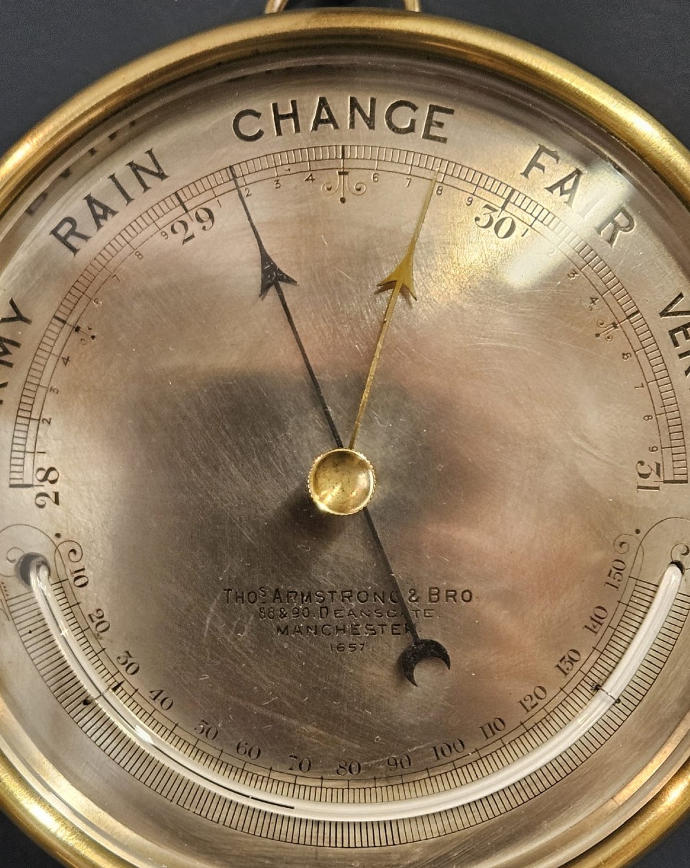 A circular Victorian brass aneroid barometer featuring an engraved silvered dial with weather scale, integrated thermometer, and a top suspension ring. View VI