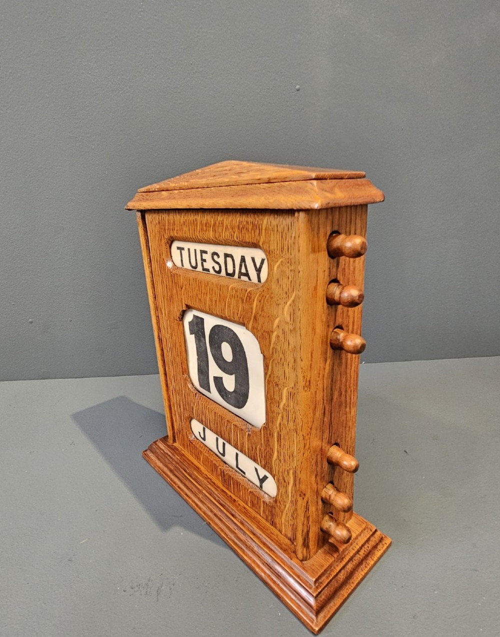 A late Victorian figured oak perpetual desk calendar with turned side knobs and three apertures displaying the day, date, and month. View VI