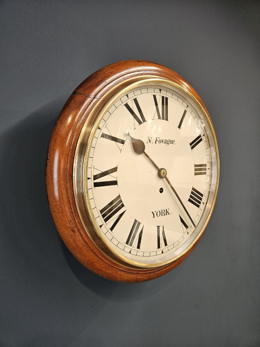 A round late Victorian mahogany wall clock featuring a brass bezel, white painted dial with black Roman numerals, and signature reading N. Fovague York. View II