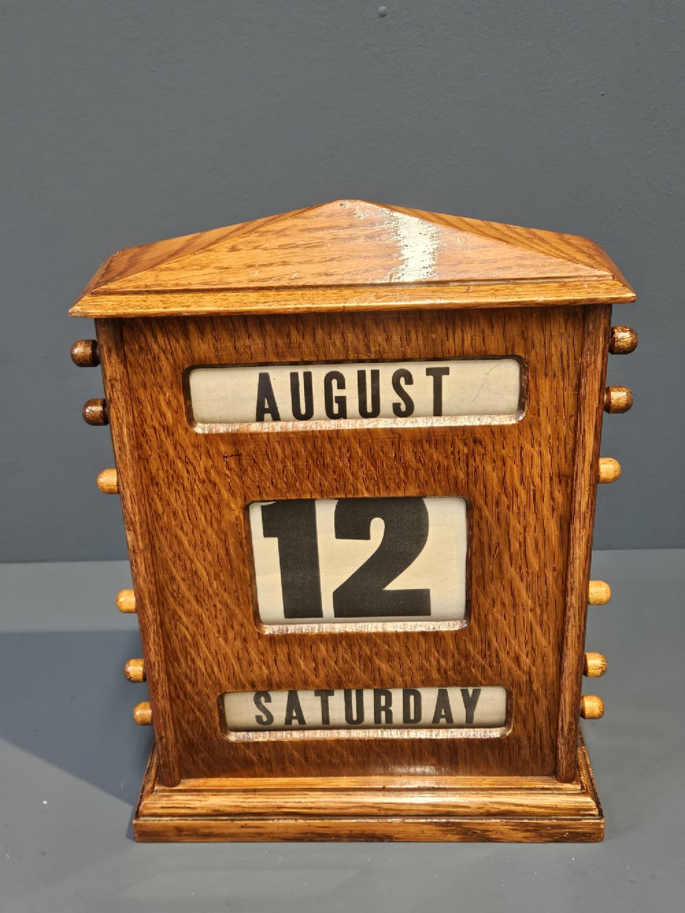 An Edwardian oak perpetual desk calendar featuring side turning knobs, apertures displaying printed day, date, and month rolls, and a gabled pediment top. View II