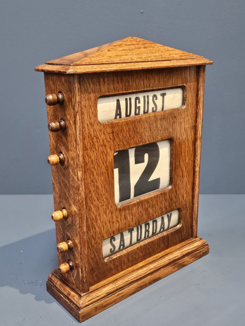 An Edwardian oak perpetual desk calendar featuring side turning knobs, apertures displaying printed day, date, and month rolls, and a gabled pediment top. View V