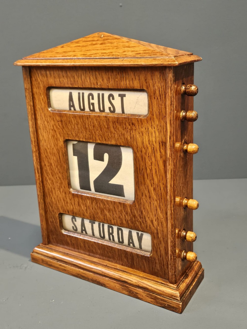An Edwardian oak perpetual desk calendar featuring side turning knobs, apertures displaying printed day, date, and month rolls, and a gabled pediment top. View VI