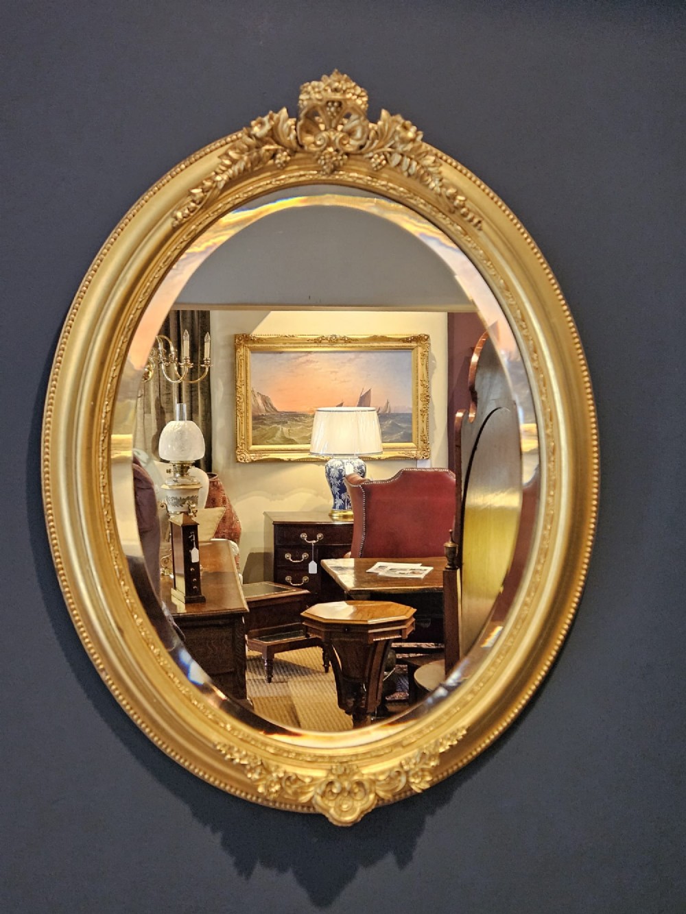 A mid-Victorian oval wall mirror featuring a giltwood frame adorned with carved cresting, beaded borders, and a bevelled mirror plate.