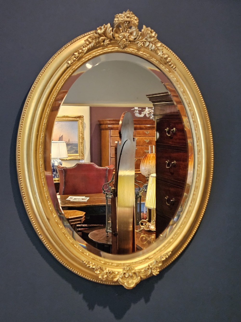 A mid-Victorian oval wall mirror featuring a giltwood frame adorned with carved cresting, beaded borders, and a bevelled mirror plate. View II