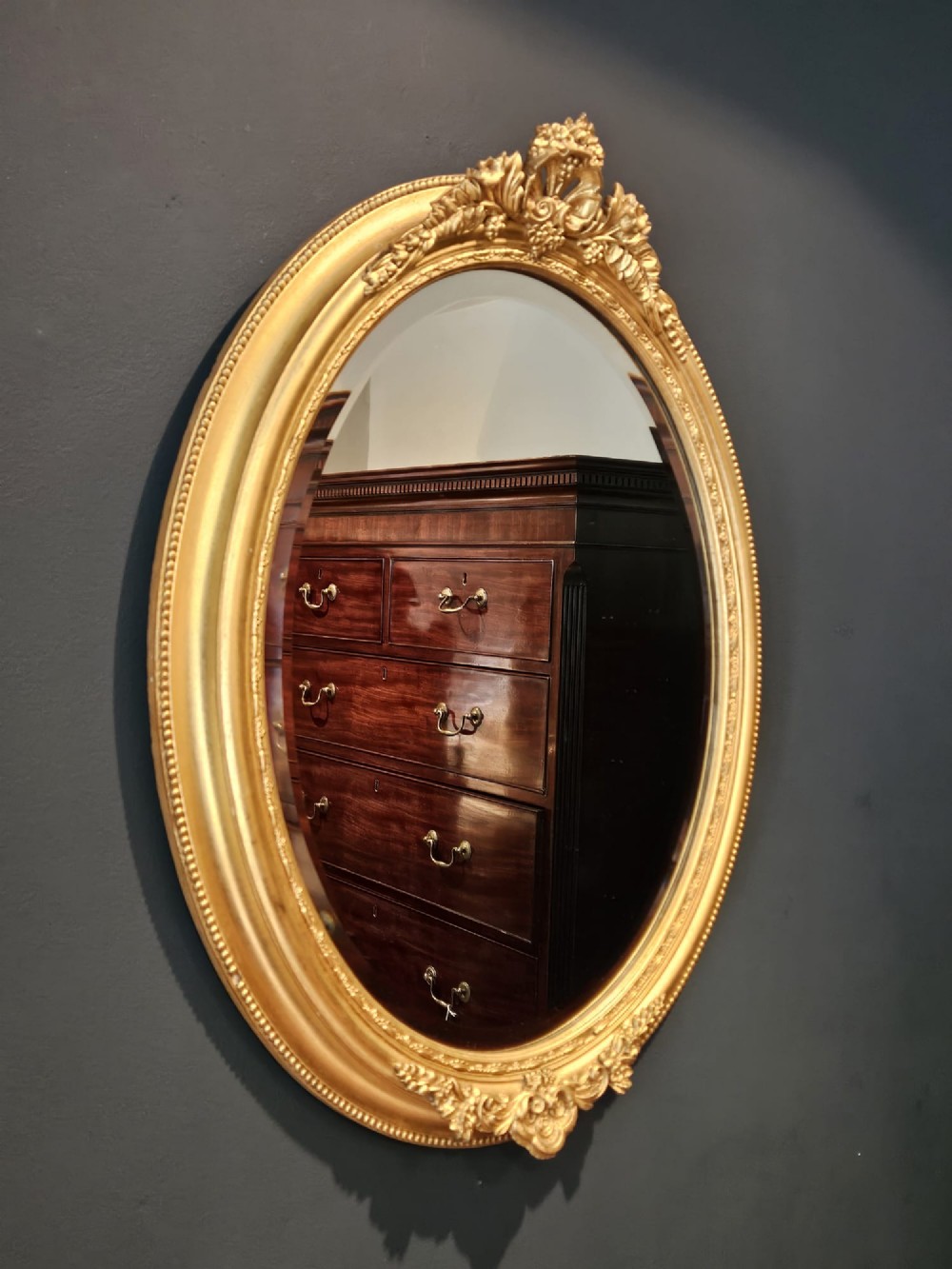 A mid-Victorian oval wall mirror featuring a giltwood frame adorned with carved cresting, beaded borders, and a bevelled mirror plate. View V