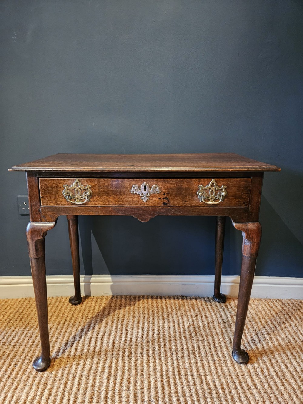 A George II dark oak side table with a single drawer, pierced brass handles, a shaped apron, and four cabriole legs terminating in pad feet.