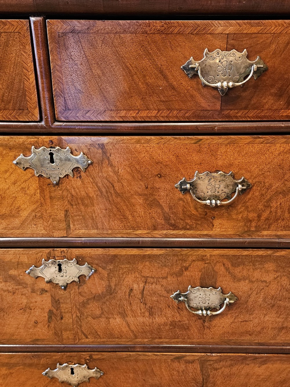 A George I walnut chest of drawers featuring herringbone crossbanding, brass engraved hardware, and bun feet against a dark wall. View II