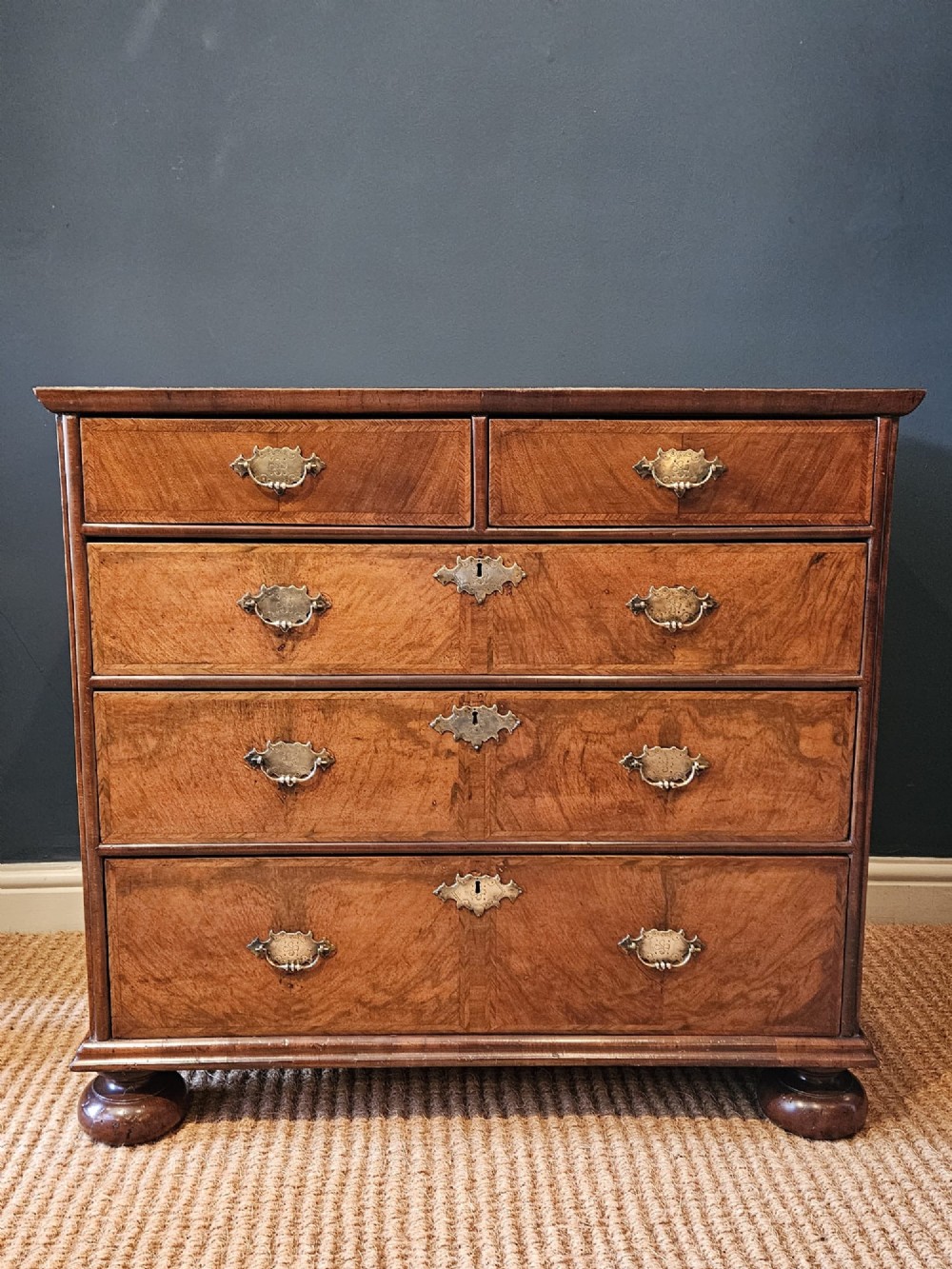 A George I walnut chest of drawers featuring herringbone crossbanding, brass engraved hardware, and bun feet against a dark wall. View III