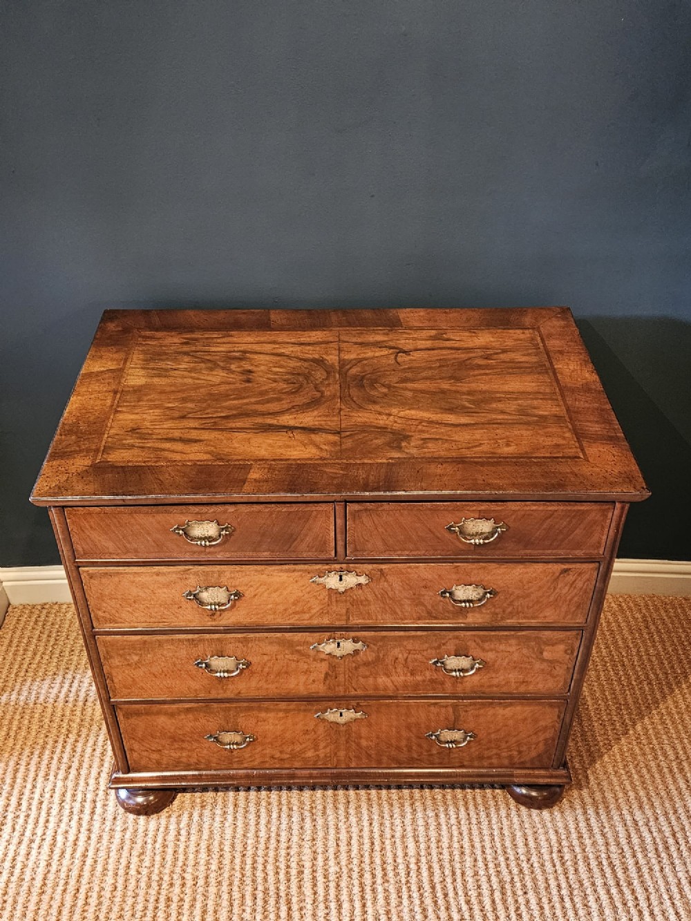 A George I walnut chest of drawers featuring herringbone crossbanding, brass engraved hardware, and bun feet against a dark wall. View IV